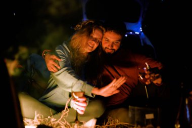 Bir çift bir minibüste oturup şöminenin önünde sıcak bir anı paylaşıyorlar. Doğanın güzelliği ve parıldayan ışıklarla çevriliyken gülüp, içkilerini yudumluyorlar..