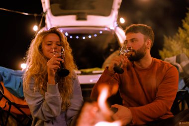 İki insan sıcak bir kamp ateşinde rahatlar, şarap yudumlar ve birlikte eğlenirler. Atmosfer sicim lambaları ve arka planda bir minibüsle rahat..