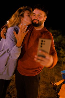 Bir çift, gece kamp yaparken mutlu bir şekilde poz veriyor. Sıcak bir gülümseme ve romantik bir öpücük. Atmosfer, yakındaki bir kamp ateşiyle aydınlanıyor..