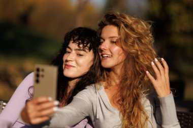 İki genç kadın birbirine yakın oturuyor ve parkta selfie çekiyorlar. Parlak gülümsemelerle, yemyeşil ve sıcak güneş ışığıyla çevrili güneşli bir günün tadını çıkarırlar..
