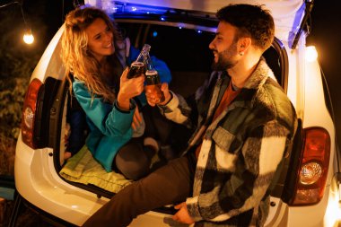 Genç bir çift arabalarının bagajında oturmuş, içki içerek kutluyorlar. Dışarıdaki ortam geceleri rahat ve samimi bir kamp ortamı yaratıyor..