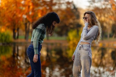 İki arkadaş, sakin bir göletin yanında neşeli bir anı paylaşıyor. Parlak sonbahar renklerinin ve huzurlu bir park ortamının arasında gülüyor ve etkileşime geçiyorlar..