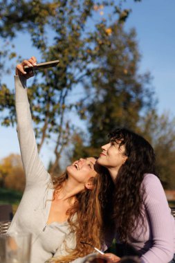 İki arkadaş, etraflarında parlak güneş ışığı ve yeşillik olan bir parkta selfie çekerken mutlu bir şekilde gülümsüyor. Kahkahaları ve sevinçleri, dışarıda birlikte geçirdikleri güzel bir anı yansıtıyor..
