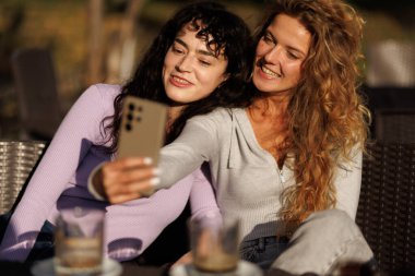 İki arkadaş bir kafede yakın oturuyorlar, akıllı telefonlarıyla selfie çekerken gülümsüyorlar. Sıcak güneş ışığı, içkilerle ve sıcak bir atmosferle çevrili neşeli anlarını güçlendirir..