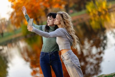 İki arkadaş, canlı sonbahar yapraklarıyla çerçevelenmiş sakin bir gölün kenarında selfie çekerken gülümsüyor. Neşeli ifadeleri mevsimin güzelliğini ve güçlü bağlarını yansıtıyor..