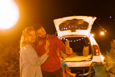 İki insan arabalarının yanında mutlu bir şekilde poz veriyor. Gece yumuşak ışıklarla doludur ve sahne, doğal bir ortamda sıcaklık ve neşe taşır..