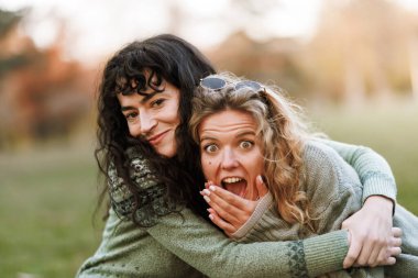 İki kadın sonbaharda bir parkta neşeli bir anı paylaşıyor. Samimi kazaklar giyiyorlar, birbirlerine eşlik etmekten ve sıcak güneş ışığından keyif alırken geniş bir gülümseme sergiliyorlar..