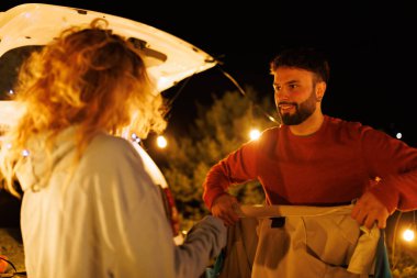 Bir çift yıldızların altında kamp yapmaya hazırlanıyor. Sıcak ışıklarla çevrili araçlarının arkasından ekipman hazırlarken gülümsüyorlar ve etkileşime geçiyorlar..