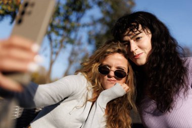 İki arkadaş bir bankta otururken eğlenceli bir anı yakalar, neşeli pozlar verir ve açık havada mavi gökyüzünün altında eğlenirler..