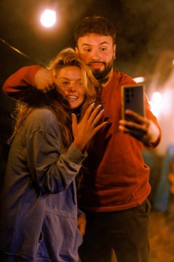 İki arkadaş gülümsüyor ve kamp ateşinin yanında sıcak bir akşamda selfie için poz veriyorlar. Ateşten gelen sıcak ışık ve dekorasyonlar mutlu bir atmosfer yaratır..