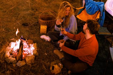 İki arkadaş sıcak bir kamp ateşinin yanında oturur, dışarıda huzurlu bir kamp alanında şarap ve atıştırmalıkların tadını çıkarırlar. Gülüşleri ve hikayeleri paylaştıkça ortam rahatlatıcı ve davetkar..