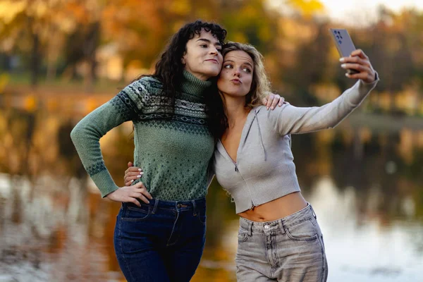 İki arkadaş sakin bir göl kenarında poz veriyor, gülümsüyor ve sıcak bir sonbahar gününün tadını çıkarıyorlar. Neşeli bir selfie çekerler, canlı yapraklarla güzel bir zemin oluştururlar..