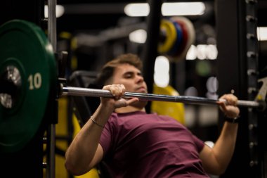Bir adam spor salonunda ağırlık kaldırırken vücuduna konsantre olarak göğüs presi egzersizi yapıyor. Spor salonunun halterinde çeşitli ekipman ve renkli plakalar var..