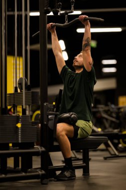 Spor salonunda oturan bir adam egzersiz malzemelerini kullanarak ağırlık kaldırmaya odaklanmış. Spor yaparken yeşil bir gömlek ve şort giyiyor..