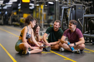 Spor salonunun zemininde oturan dört kişi bir tartışma içindeler. Onlar tableti incelerken diğer spor aletleri arka planda görünür..