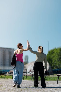 Parkta yüz yüze duran iki kadın. Çak bir beşlik için ellerini kaldırıyorlar. Gökyüzü açık ve arka planda ağaçlar görülebilir. Açık havada vakit geçirmekten hoşlanıyorlar..
