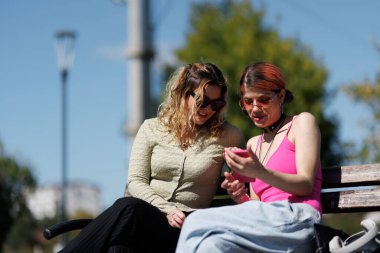 Parktaki bankta yan yana oturan iki kadın. Telefona bakıp gülüyorlar. Güneş açık bir günde parlak bir şekilde parlar. Ağaçlar çevrelerini sarmış, canlılık katıyor..