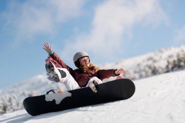 Kar kayağı yapan bir kız kayak merkezinde kar kayağı yapıyor. O geniş bir şekilde gülümsüyor ve eğleniyor gibi görünüyor. Gökyüzü açık ve arka planda dağlar görünüyor..