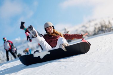 Bir kız tek bacağı havada kameraya gülümseyen bir şekilde snowboard 'unda oturuyor. Diğer snowboardcular arka planda, dağ yamacındaki karlı günün tadını çıkarırken görülebilir..
