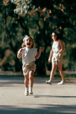 Mutlu bir çocuk gündüz vakti parkta koşarken bir yetişkinin durup izlediği, kaygısız bir neşe ve açık hava aktivitesi yakaladığı görülüyor..