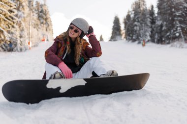 Bir kız kış manzarasında snowboard 'a ara veriyor. Kask ve eldiven takıyor ve bir kayak merkezinde karla kaplı zeminde dinlenirken mutlu görünüyor..
