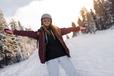 Karlı bir yamaçta kolları açık duran bir kız. Kask takıyor ve güneş gözlüğü takıyor. Güneş arka planda ışıl ışıl parlıyor. Ağaçların ve karın kış manzarasını aydınlatıyor..