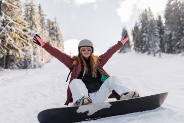 Karlı bir bölgede kollarını kaldırmış bir kız snowboard 'una oturuyor. Bir kask ve parlak eldivenler giyiyor. Etrafını ağaçlar sarmış ve diğer insanlar arka planda karın tadını çıkarırken görülebiliyor..