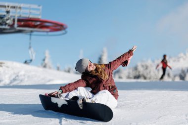 Kar kayağı yaparken bir kız oturur, gülümser ve kollarını uzatır. Arka planda, diğer insanlar kayak yapıyor ve yamaçlarda kış havasının tadını çıkarıyorlar..