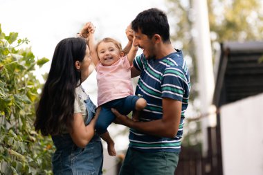 Neşeli bir aile arka bahçelerinde birlikte kaliteli zaman geçirir. Küçük çocuklarını kaldırırken, mutluluğu ve bağlantıyı teşvik ederken kahkahalarla bağlandıkları anların tadını çıkarıyorlar..