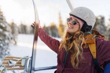 Heyecandan kolları kalkmış bir kız, kayak asansörüne biniyor. Güneş gözlüğü ve kask takıyor. Bölgenin etrafında karla kaplı ağaçlar var. Kış boyunca aydınlık ve güneşlidir..