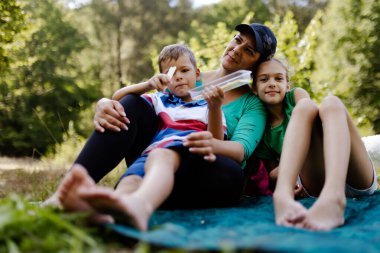 Bir anne ve iki çocuğunun, doğa ile çevrili bir battaniyenin üzerinde oturup birlikte olmanın ve boş zamanların tadını çıkardıkları rahatlatıcı bir an..
