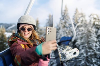 Kız fotoğraf çekmek için akıllı telefon tutarken kayak asansöründe. Kask takıyor ve kış günü etrafı karla kaplı ağaçlarla çevrili..