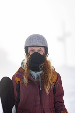 Karlı bir bölgede kask ve siyah bir maske takmış bir kız duruyor. Bir elinde snowboard tutuyor. Sahne soğuk ve karlı, kar yağışı nedeniyle görüş mesafesi düşük..