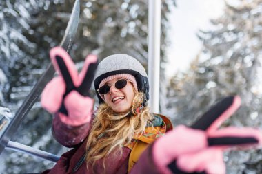 Bir kız, iki eliyle barış işaretleri göstererek, kayak asansöründe zaman geçirmekten zevk alır. Kışın arka planda kar ve ağaçlarla çevrili bir kask ve güneş gözlüğü takıyor..