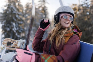 Bir kız karlı ağaçlarla çevrili bir teleferiğe binmekten hoşlanır. Gülümserken ve el sallarken kask ve renkli eldivenler takıyor. Sahne berrak gökyüzü ile aydınlık.