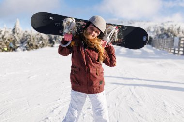 Kar üzerinde omuzlarında snowboard 'uyla duran bir kız. Karla kaplı ağaçlar ve açık bir gökyüzü görülebilir. Kış sporu sahnesi için sıkı giyinmiş..