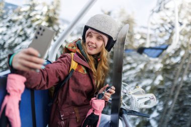 Bir kız dağlarda kayak asansöründe. Telefonunu tutuyor ve selfie çekerken gülümsüyor. Etrafındaki ağaçları kar kaplıyor. Kask ve kayak kıyafeti giyiyor, kayak yapmaya hazır..