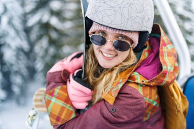 Yüzünde güneş gözlüğü ve miğfer olan bir kız asansörde oturuyor. Kış güneşinde arkadaşlarıyla karlı bir dağda kayak yapmaya hazırlanırken elinde bir snowboard tutuyor..