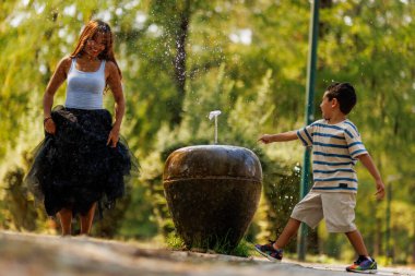 Bir anne ve oğlu, güneşli bir günde yemyeşil bir parkta dekoratif bir fıskiyenin yanında oynarken su sıçratmaktan ve gülmekten zevk alırlar..