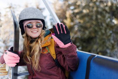 Kız dağ eteklerinde kayakla gezmekten hoşlanıyor. Kask ve eldiven takıyor, gülümsüyor ve el sallıyor. Kar zemini kaplıyor ve etrafındaki ağaçlar gün ışığında.