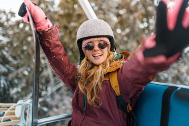 Bir kız büyük bir gülümsemeyle kayak asansörüne biniyor. Kask takıyor ve güneş gözlüğü takıyor. Karla kaplı ağaçlar asansörün etrafını sarar ve güneş sahnede parlar..