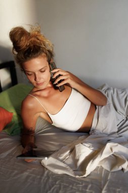 Bir kadın rahat bir şekilde bir yatağa uzanıyor, akıllı telefonunu kullanırken bir telefon görüşmesi yapıyor. Güneş ışığı rahat odayı aydınlatır, rahat atmosferi güçlendirir..