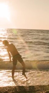 A young boys silhouette is captured as he playfully throws rocks into the calm ocean waves at sunset, with golden sunlight reflecting beautifully on the water during a peaceful summer evening on the