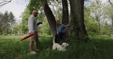 Çiftler, köpekleriyle birlikte doğada rahatlatıcı bir pikniğin tadını çıkarırlar. Büyük bir ağacın altında huzurlu bir anı paylaşıyorlar, beraberliği teşvik ediyorlar ve açık havaya karşı sevgi besliyorlar..