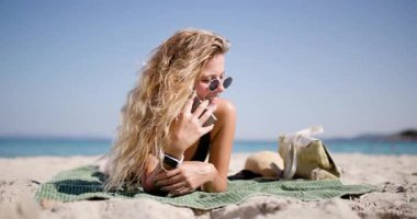 Bir kişi plaj havlusunda uzanmış güneşlenirken telefonla konuşuyor. Okyanus dalgaları yavaşça arka plana çarpar ve barışçıl bir atmosfer yaratır..