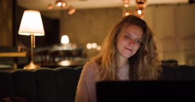Akşamları sıcak, davetkar bir restoran atmosferinde verimli bir oturumdan sonra laptopunu kapatırken uzun saçlı bir kız gülümsüyor. Yumuşak ışıklandırma havaya renk katar..