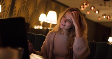 Şık bir restoran ortamında, uzun saçlı bir kız laptopuna odaklanmış, sigara içerken özenle çalışıyor. Sıcak ışıklar rahat bir ortam yaratıyor, gece görevleri için mükemmel..