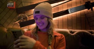 Turuncu kazaklı ve bereli gülümseyen sarışın kadın kayak merkezinde deri koltukta akıllı telefon kullanıyor. Kayaktan sonra mutlu ve rahatlamış..