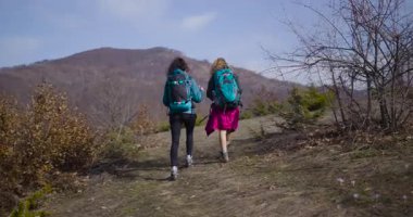 İki bayan arkadaş toprak bir yolda dağ yürüyüşünden zevk alıyor. Sırt çantaları taşıyorlar ve manzaranın ve temiz havanın tadını çıkarıyorlar..