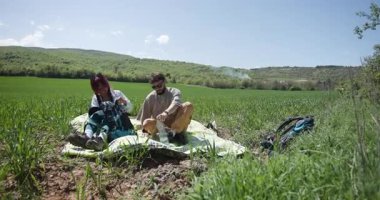 Genç bir çift yeşil bir alanda rahatlatıcı bir pikniğin tadını çıkarıyor. Su içiyorlar, sohbet ediyorlar ve etraflarındaki güneş ışığını ve doğayı emiyorlar..